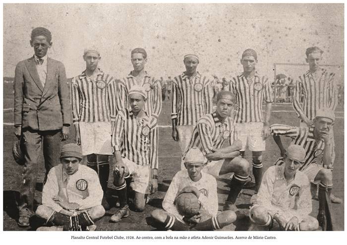  4 a.  Foto em preto e branco, time do Planalto Central Futebol Clube 1928 Os jogadores trajam camisas de mangas compridas listradas, shorts e chuteiras. Ao centro com a bola na mão o atleta Ademir Guimarães, ele é de pele clara, ssua um gorro. Ao lado esquerdo, um homem próximo ao jogador segura um chapéu na mão direita. Realização: Academia Planaltinense de Letras, Artes e Ciências (APLAC), pelo Ecomuseu Pedra Fundamental e pelo Coletivo Nativo Audiodescrição produzida  pelo Instituto de Promoção das Pessoas com Deficiência Visual Audiodescritora: Elma Lúcia Rodrigues Consultor: Fernando Rodrigues Este projeto é promovido com recursos do Fundo de Apoio a Cultura do DF